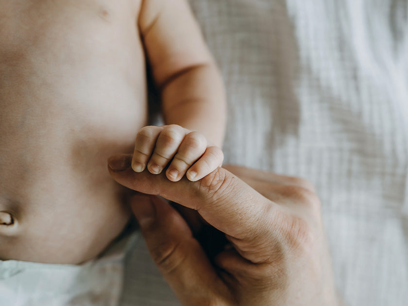 Main d’un nouveau-né serrant le doigt d’un adulte, sur un fond de drap blanc.