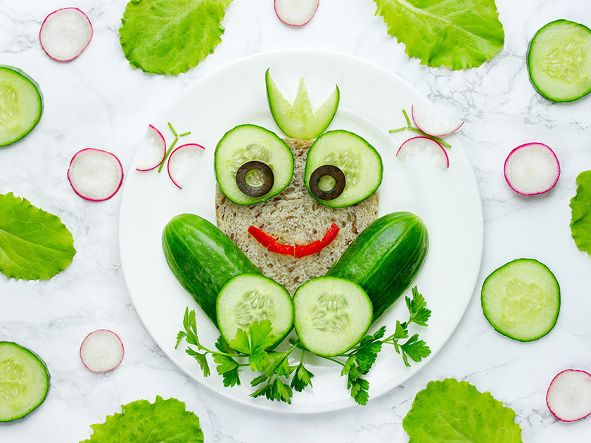 Assiette avec une composition alimentaire en forme de grenouille souriante, réalisée avec des concombres, olives, radis, salade et pain, sur fond de table marbrée.