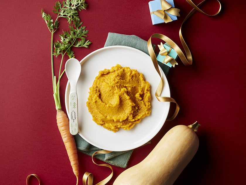 Assiette de purée de légumes orange servie avec une cuillère pour bébé, accompagnée d’une carotte, d’une courge butternut et de décorations colorées, sur un fond rouge.