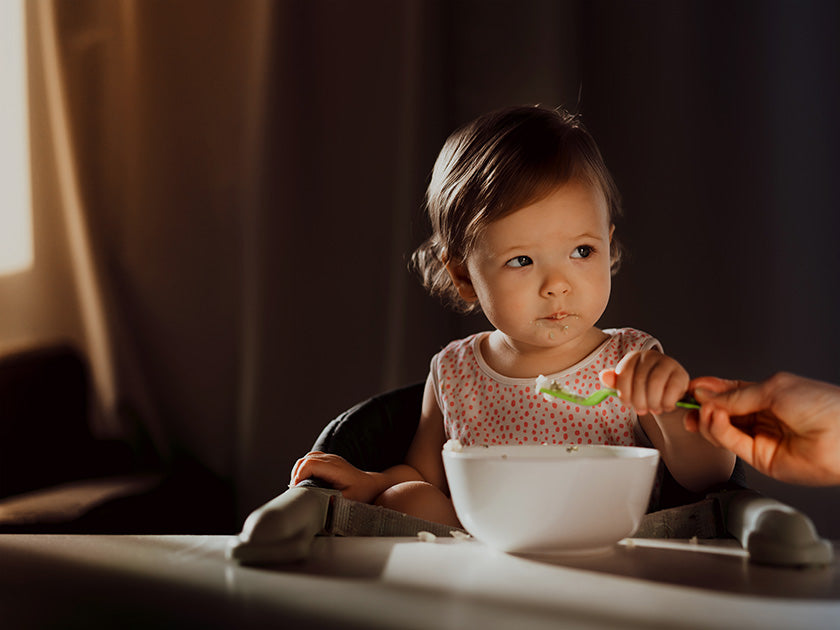 Petite fille assise dans une chaise haute, regardant sur le côté pendant qu'un adulte lui tend une cuillère de nourriture. Elle a un bavoir rose à pois et un bol blanc posé devant elle.