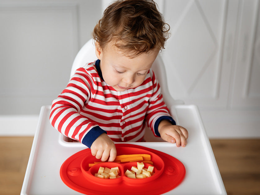 Jeune enfant assis dans une chaise haute, portant un pyjama rayé rouge et blanc, en train de manger des morceaux de fruits et de légumes dans une assiette rouge compartimentée