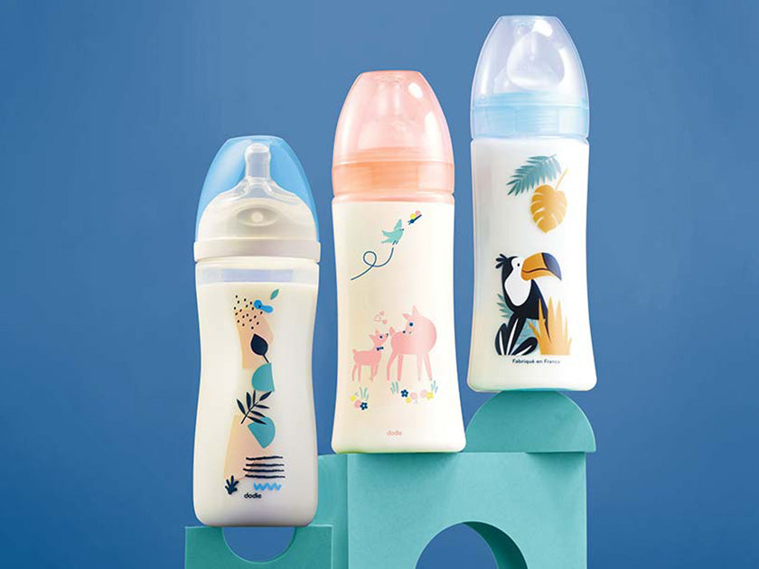 Trois biberons pour bébé aux motifs colorés (animaux et plantes), posés sur des blocs bleus de la marque Dodie, avec un fond bleu uni.