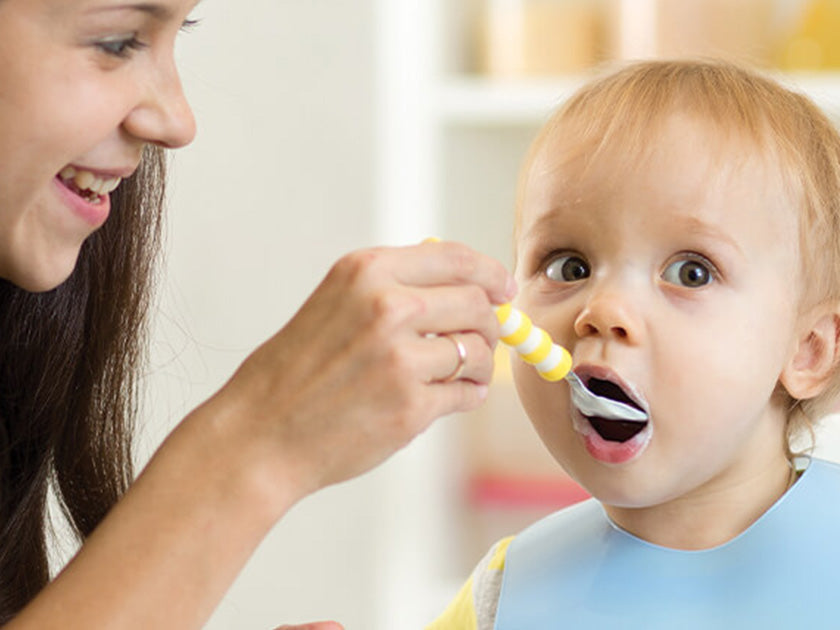 Une femme souriante donne à manger à un enfant avec une cuillère jaune et blanche. L’enfant porte un bavoir bleu et regarde vers l’avant.