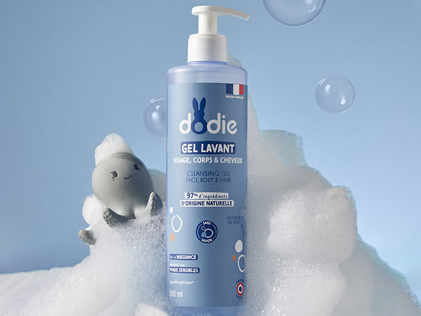 Flacon de gel lavant Dodie pour visage, corps et cheveux, posé sur une surface avec mousse blanche et un petit jouet éléphant gris, fond bleu clair.