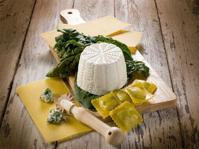 Planche en bois posée sur une table rustique, garnie de feuilles d’épinards, d’un fromage de ricotta, de raviolis farcis, de pâte fraîche et d’un petit ustensile en bois.