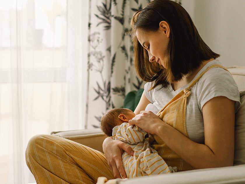 Mère assise dans un fauteuil, allaitant tendrement son bébé emmitouflé dans une gigoteuse rayée, dans une pièce lumineuse et chaleureuse.