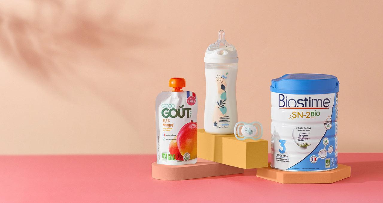 Gourde de compote bio à la mangue Good Goût, biberon décoré avec tétine, sucette pour bébé, et pot de lait infantile en poudre Biostime SN-2 Bio Plus, tous posés sur des socles colorés dans un décor pastel.
