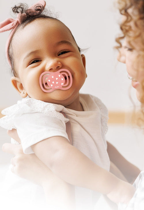 Une mère tenant son enfant dans les bras ; l’enfant utilise une tétine rose Dodie .