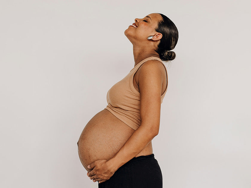 Femme enceinte debout de profil, souriante et les yeux fermés, tenant son ventre arrondi avec tendresse.