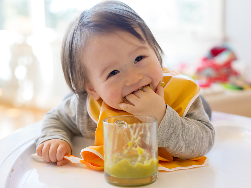 Bébé souriant portant un bavoir orange, mangeant avec les doigts depuis un pot de purée verte posé sur une table.