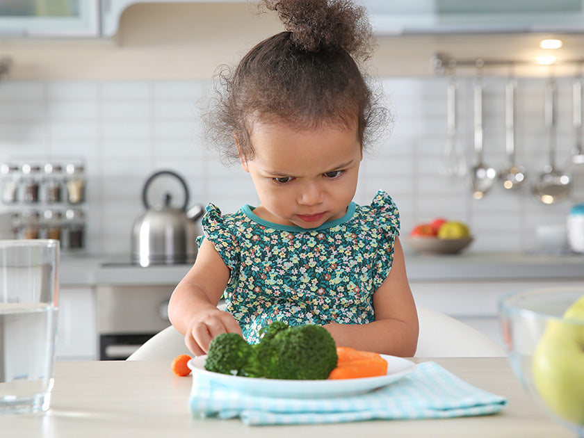 Une petite fille fronce les sourcils en regardant une assiette de légumes, notamment du brocoli et des carottes, dans une cuisine moderne.