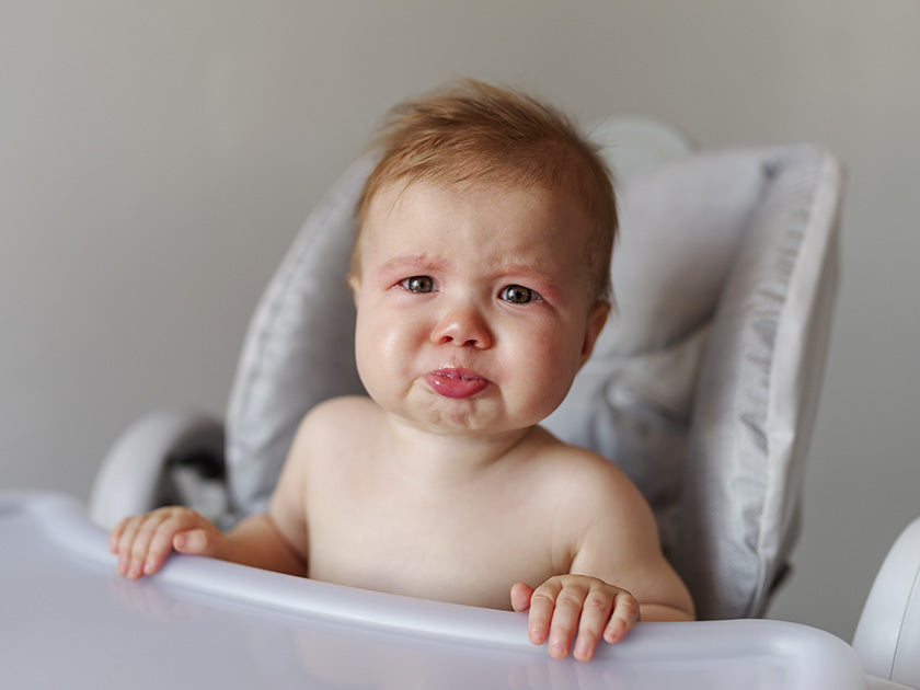 Bébé pleurant assis dans une chaise haute, regard triste, fond clair.