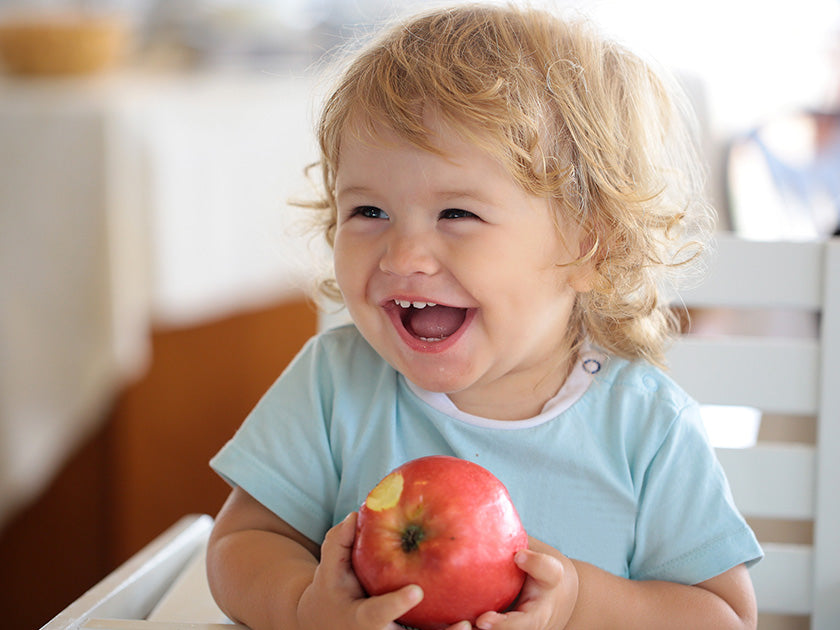 Un jeune enfant aux cheveux bouclés blonds sourit joyeusement en tenant une grosse pomme rouge, assis sur une chaise haute dans une pièce lumineuse.