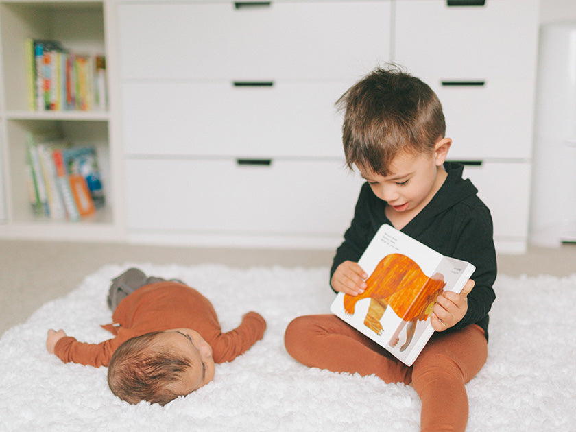 Jeune enfant assis sur un tapis blanc, lisant un livre illustré à un bébé allongé à côté de lui dans une chambre.