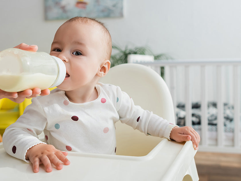 Un bébé en pyjama blanc à pois colorés est assis dans une chaise haute et boit au biberon tenu par un adulte