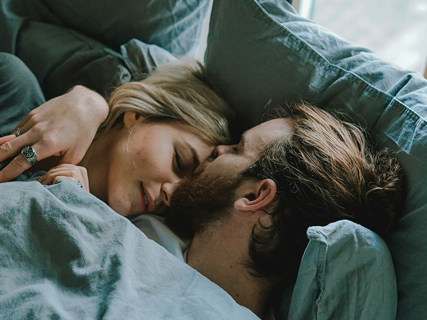Couple allongé dans un lit, tendrement enlacé sous une couette bleue, partageant un moment d'affection. 
