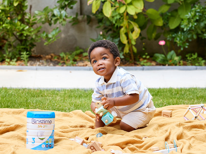 Jeune enfant jouant avec des jouets en bois sur une couverture jaune dans un jardin, avec un paquet de lait Biostime à côté.