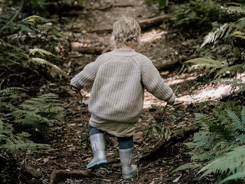 Un jeune enfant portant un pull en laine beige et des bottes de pluie bleues marche sur un sentier forestier entouré de fougères.