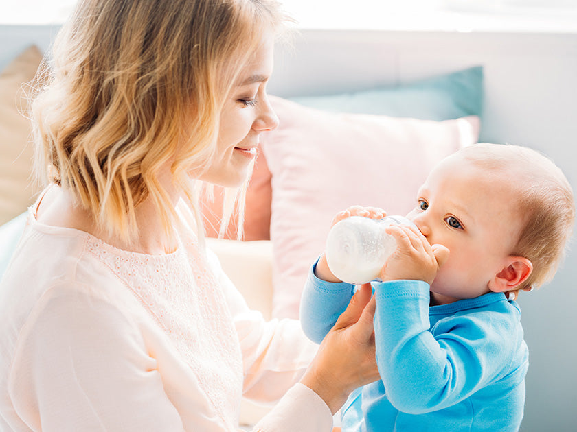 Une femme souriante aide un bébé vêtu de bleu à boire au biberon dans un cadre intérieur lumineux et chaleureux.