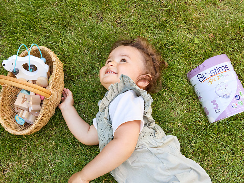 Bébé allongé sur l'herbe, souriant, à côté d’un panier de jouets et d’une boîte de lait infantile Biostime.