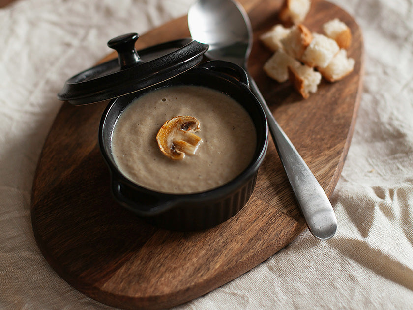 Petit pot noir avec couvercle posé sur une planche en bois, contenant une soupe ou crème de champignons, garnie d'une tranche de champignon. 