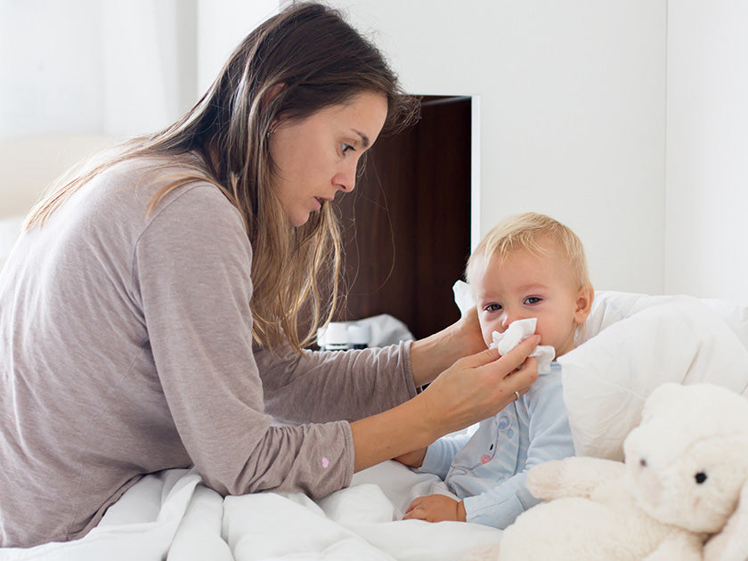 Maman essuie le nez de son bébé assis dans un lit, avec un doudou à côté