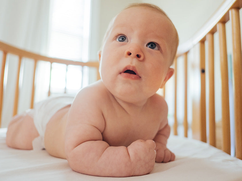Bébé allongé sur le ventre dans un lit à barreaux en bois, regardant vers le haut avec curiosité. Il porte uniquement une couche et semble éveillé et attentif. La chambre est lumineuse avec des murs clairs.