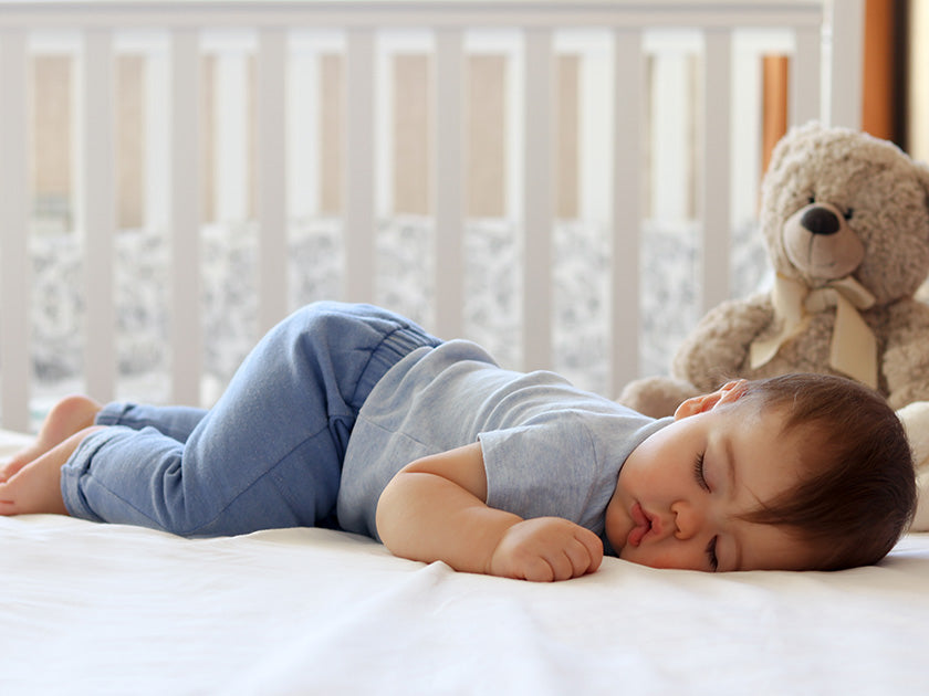 Bébé dormant paisiblement sur le côté dans un lit blanc, avec un ours en peluche en arrière-plan.