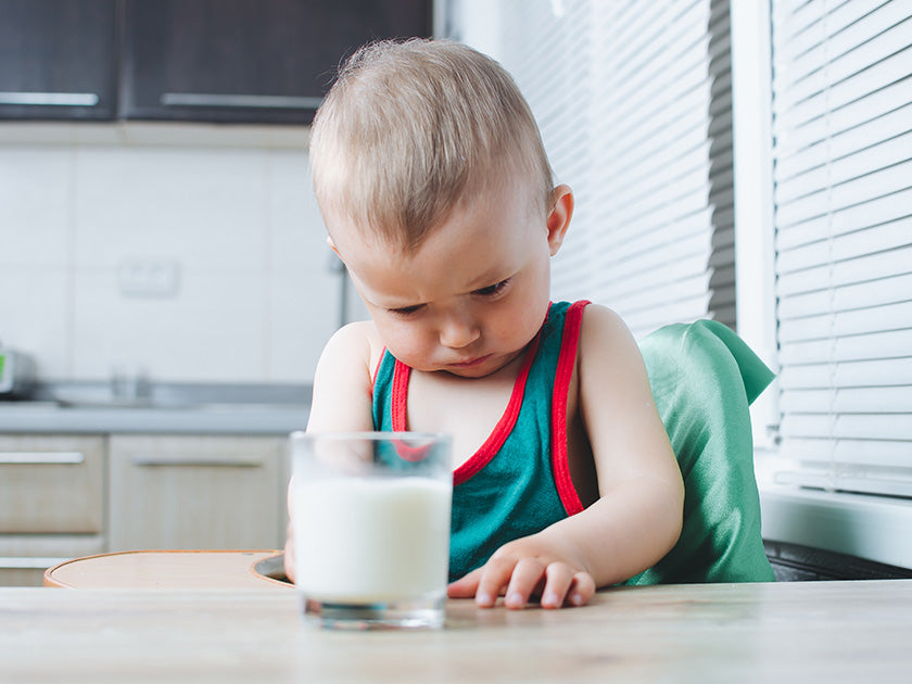  Jeune enfant assis à une table dans une cuisine, regardant d’un air concentré ou mécontent un verre de lait posé devant lui.