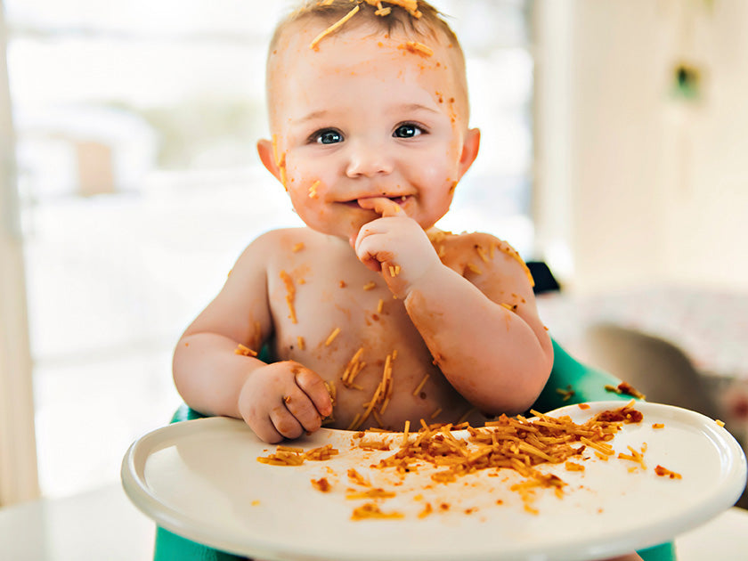 Bébé souriant assis dans une chaise haute, couvert de sauce et de morceaux de nourriture, jouant avec des spaghettis sur la tablette.