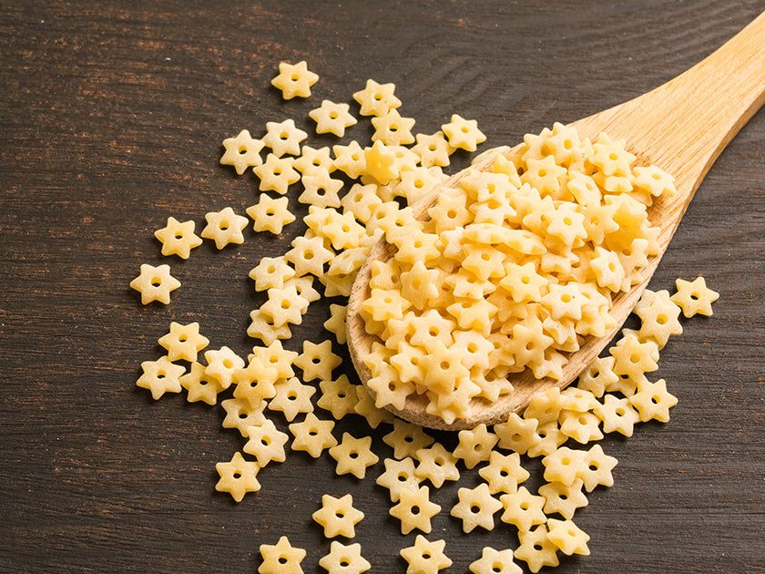 Petites pâtes en forme d’étoiles, présentées sur une cuillère en bois et dispersées sur une surface en bois foncé.
