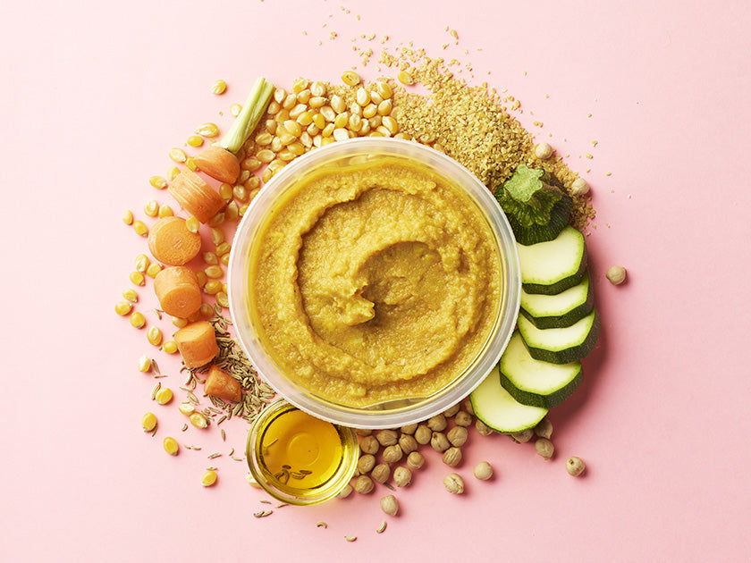 Bol de houmous crémeux entouré d’ingrédients frais sur fond rose : pois chiches, rondelles de courgette, carottes, brocoli, graines de sésame et un petit récipient d’huile d’olive