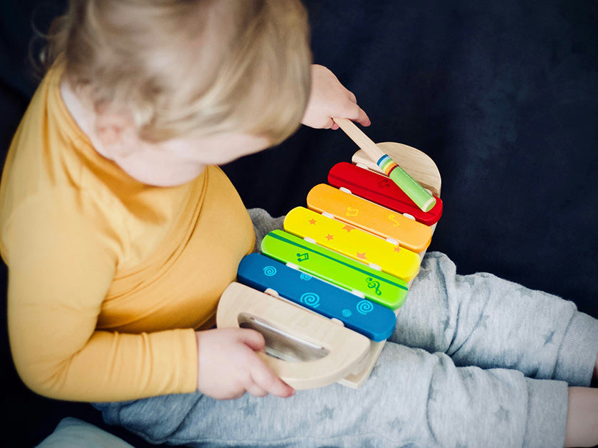 Jeune enfant jouant du xylophone coloré avec une baguette, assis sur un fond sombre.
