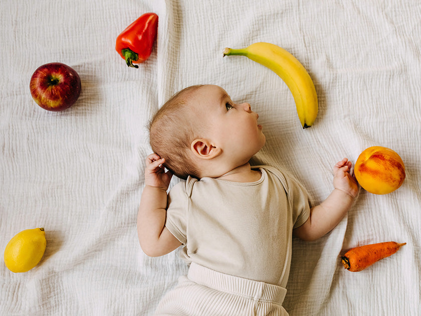 Un bébé allongé sur un tissu clair, regardant vers la droite, entouré de divers fruits et légumes colorés, dont une pomme, un poivron rouge, une banane, une pêche, un citron et une carotte.