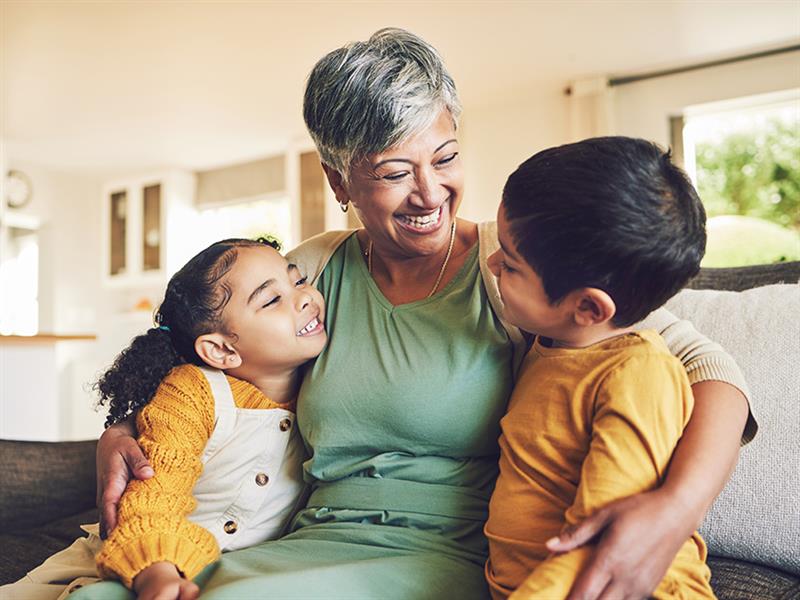 Une femme âgée assise sur un canapé, souriant et étreignant deux jeunes enfants, un garçon et une fille, dans un moment de tendresse et de joie familiale.