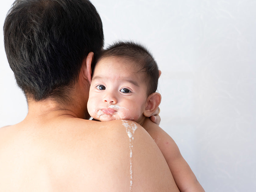 Bébé regardant vers l’avant, avec un peu de nourriture sur le visage, tenu par un adulte vu de dos.