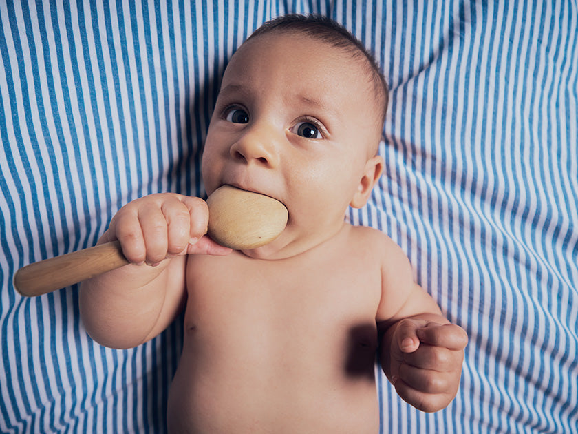 Bébé allongé sur un drap rayé bleu et blanc, tenant une grande cuillère en bois dans sa bouche.