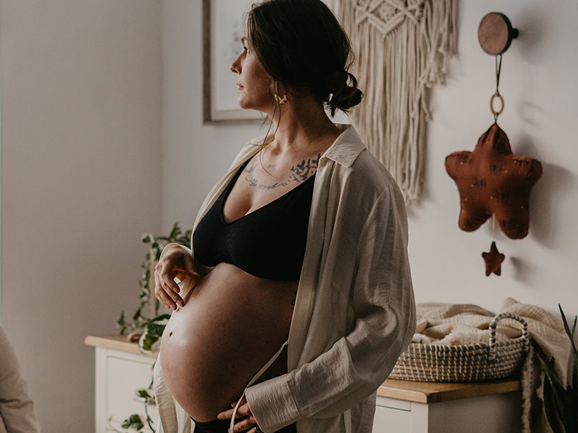 Femme enceinte en robe légère ouverte, portant un soutien-gorge noir, regardant sur le côté dans une pièce décorée avec un mobile étoile et un panier en osier.