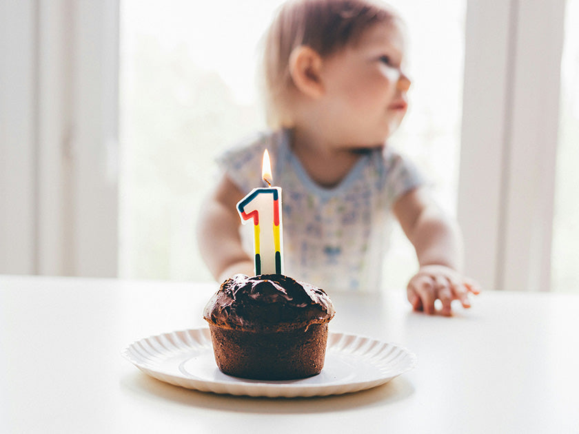Un bébé est assis près d’un petit gâteau au chocolat avec une bougie en forme de chiffre 1 allumée, marquant son premier anniversaire.