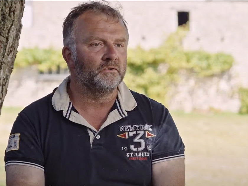 Capture d'écran d'une vidéo intitulée "Biostime - 24h dans la vie de David, agriculteur", montrant un homme barbu portant un polo bleu, assis à l'extérieur près d'un arbre, avec un bâtiment en arrière-plan.