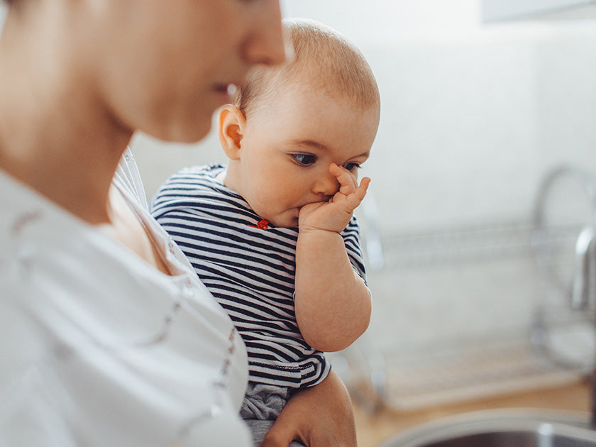 Mère tenant son bébé habillé d’un body rayé, le bébé portant son pouce à la bouche, dans une cuisine lumineuse.