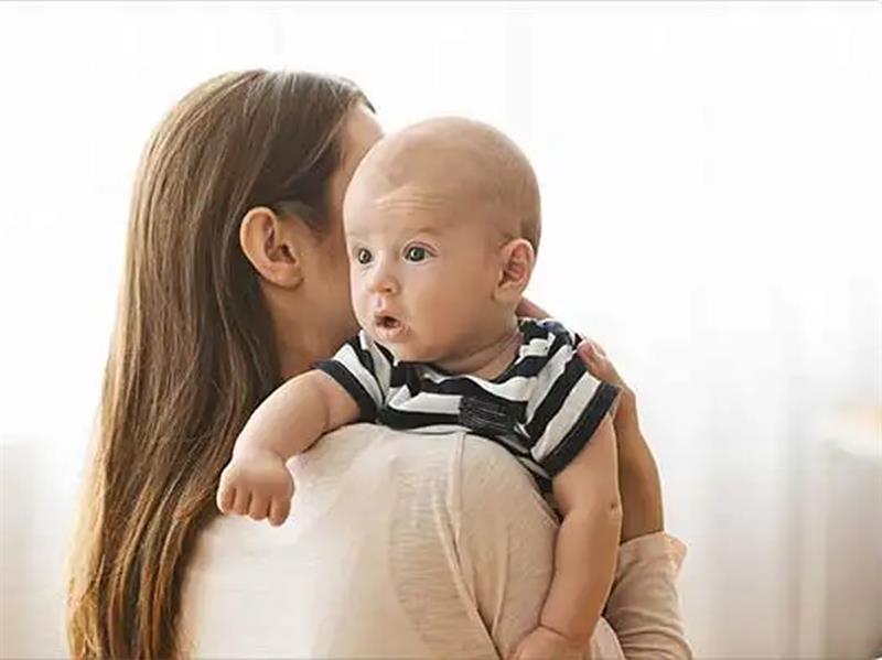 Bébé porté contre l’épaule d’une femme de dos, regardant autour de lui avec curiosité.