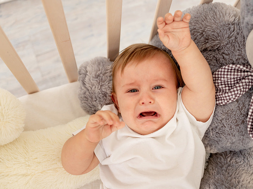 Bébé en pyjama blanc dans un lit, en train de pleurer avec un bras levé, entouré de peluches.