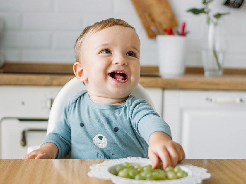 Un bébé souriant assis dans une chaise haute, tendant la main vers une assiette de raisins verts posée sur une table dans une cuisine lumineuse.