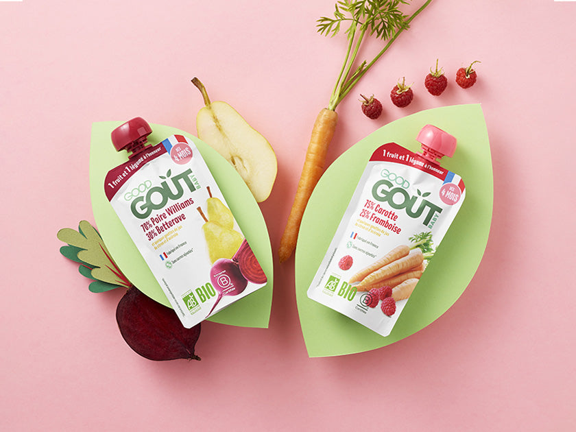 Deux gourdes de compotes bio pour bébé de la marque Good Goût, posées sur un fond rose avec des feuilles en papier vert. Autour, des légumes et fruits frais tels que poire, carotte, betterave et groseille.