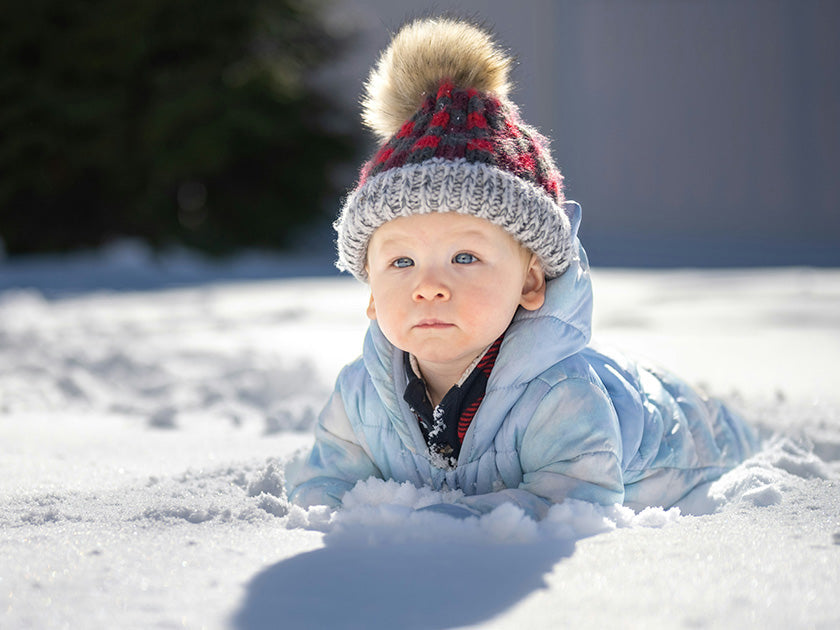 Bébé emmitouflé dans une combinaison bleue et un bonnet à pompon, allongé dans la neige par une journée ensoleillée.